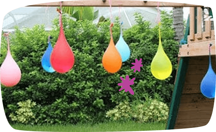 Colorful water balloons filled with water hanging from a rope in a backyard, in front of a green hedge near a wooden play structure.